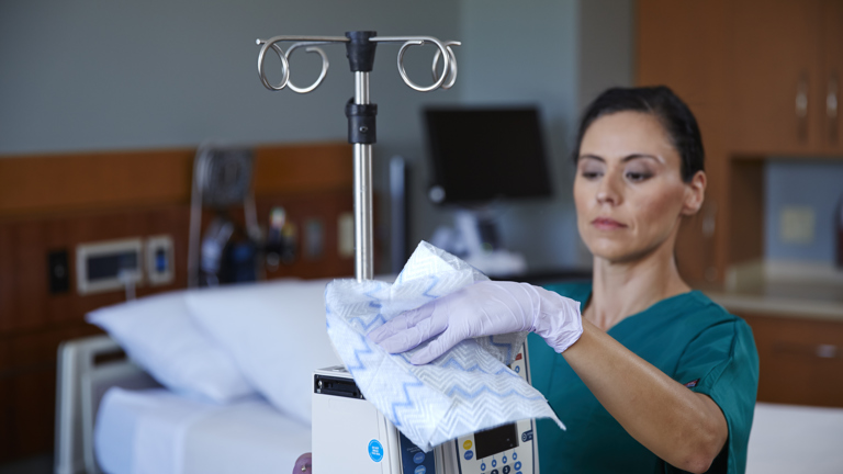 A healthcare worker wearing scrubs and gloves is shown cleaning a medical device with a wipe. The background is a hospital room with a bed.