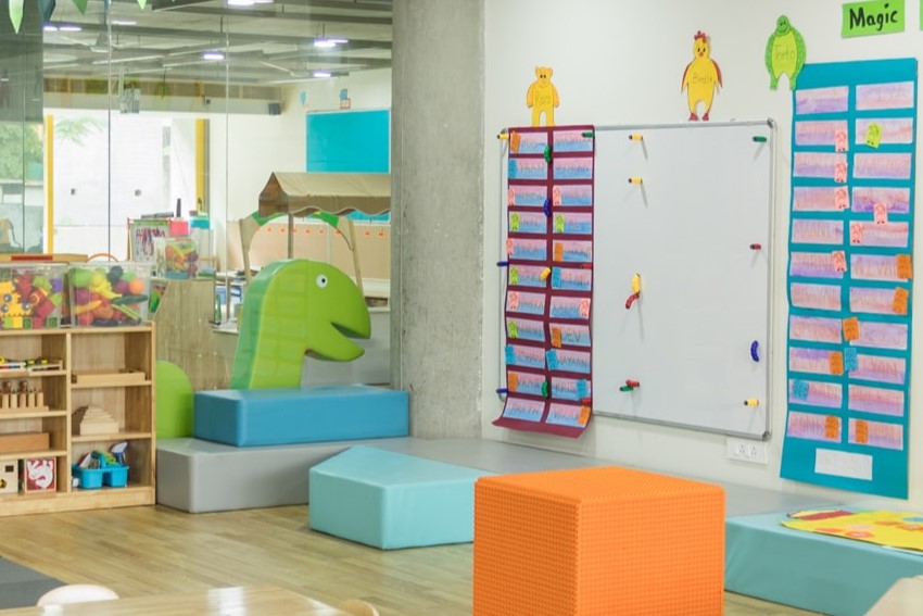 A vibrant daycare classroom with colorful toys, including a green dinosaur-shaped foam block, shelves of books and toys, and a whiteboard decorated with children's work.