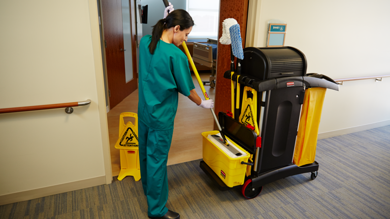 A woman in green scrubs and white gloves cleans a floor in a hospital hallway. She is pushing a yellow mop bucket and a cleaning cart. A "CAUTION Wet Floor" sign is on the floor beside her.
