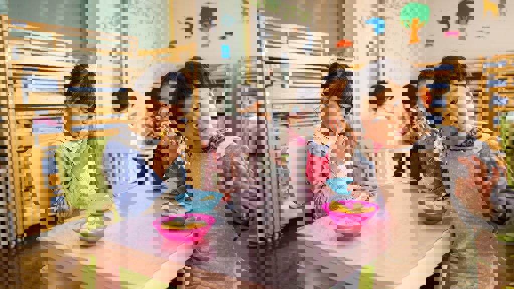 Group of young children eating snacks at a pink table, with one child in the foreground wearing an apron and looking playfully at the camera. Bunk beds are visible in the background.