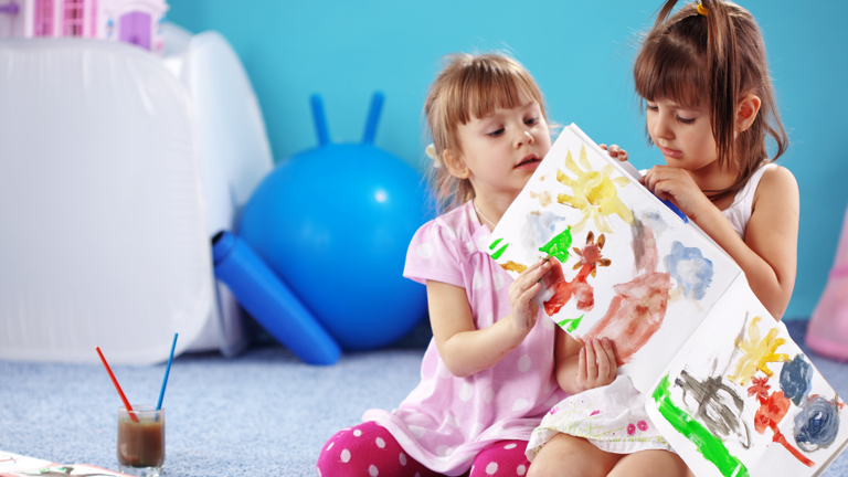 Two girls with brown hair, in pink and white dresses, sit on a gray carpet looking at a painting. A blue wall and exercise ball are in the background.