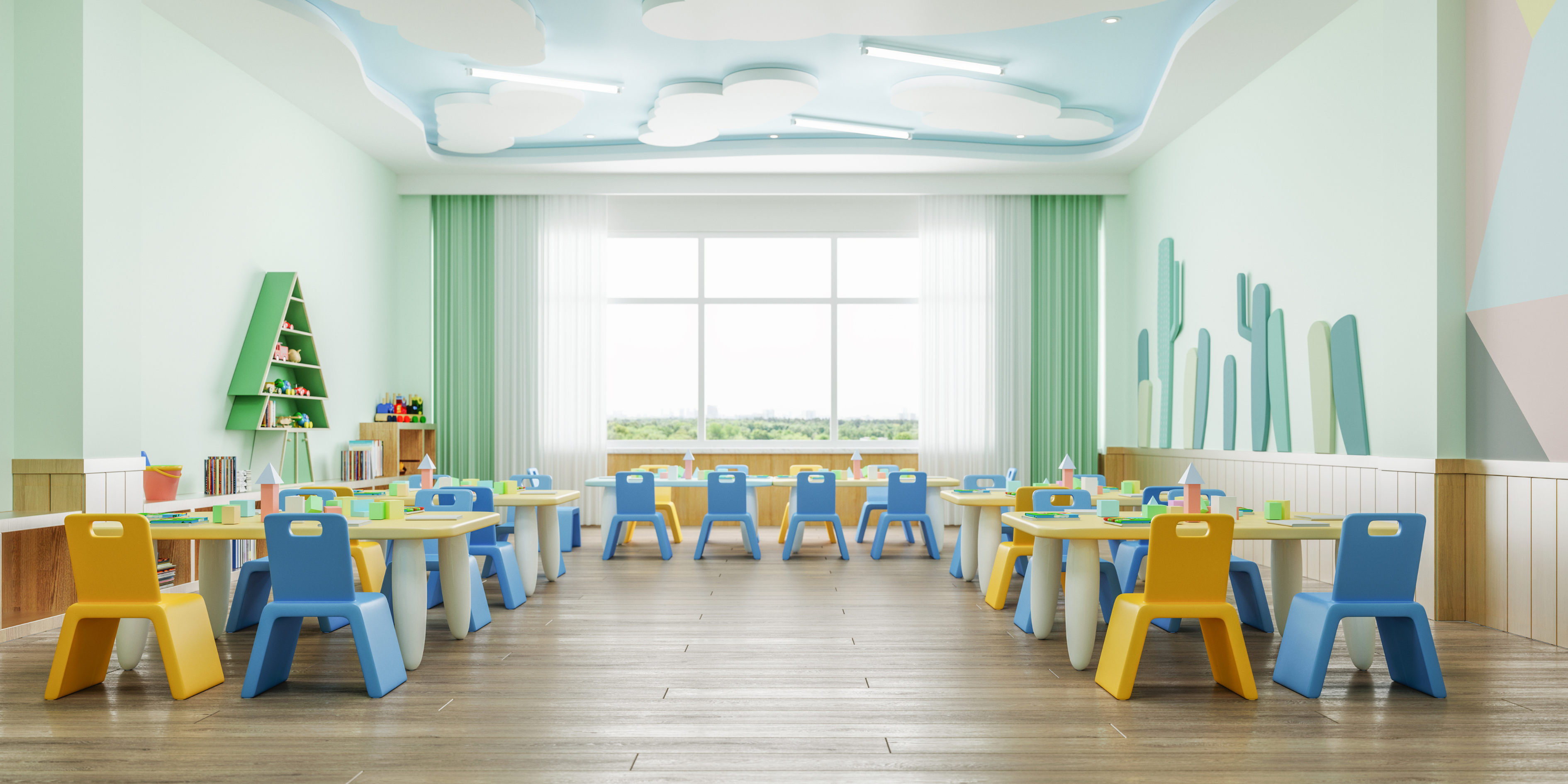 A bright, clean, and empty classroom or playroom designed for young children. The room has light green walls, a cloud-themed ceiling, and is filled with small tables and colorful yellow and blue chairs.