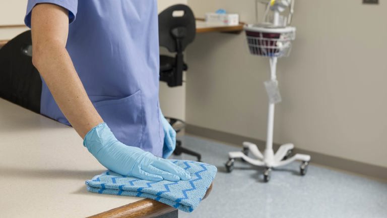 Healthcare worker cleaning a surface with a blue microfiber cloth in a hospital or clinic.