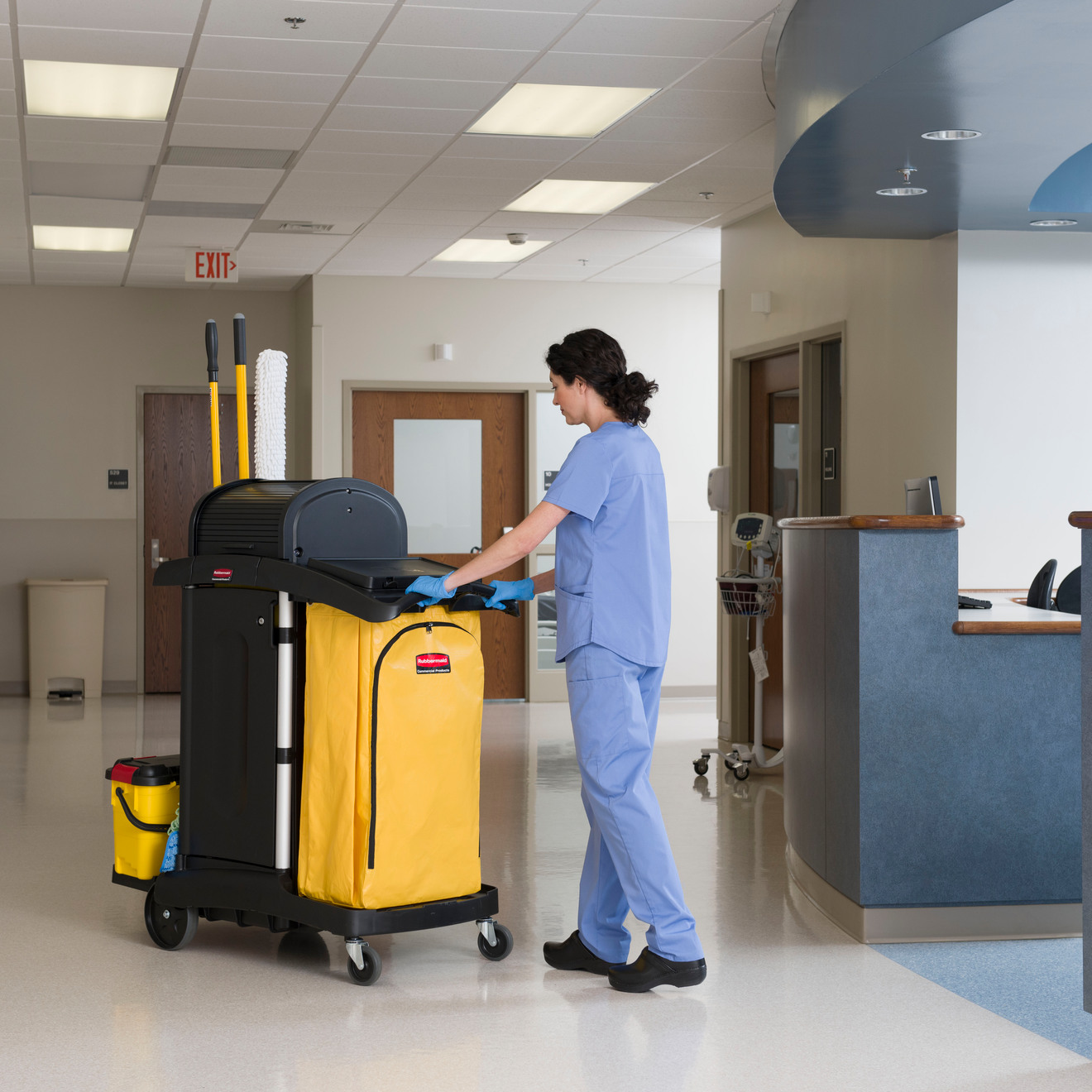 Person in blue scrubs and gloves pushing a Rubbermaid High Security Cleaning Cart with yellow bag and tools in a medical hallway.