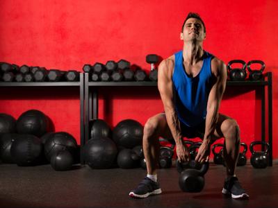 Muscular man in a blue tank top and black shorts performing a squat with kettlebells in a gym, with red wall and racks of weights in the background.