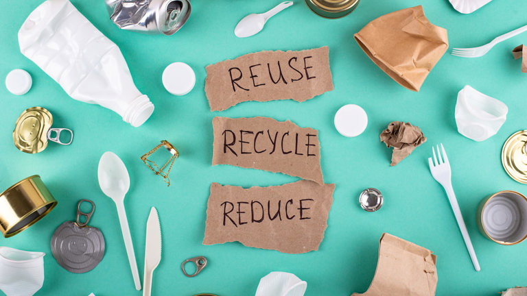 Top-down view of recyclable and non-recyclable waste—plastic bottles, aluminum and tin cans, paper, and utensils—surrounding torn cardboard labeled 'REUSE,' 'RECYCLE,' and 'REDUCE.'
