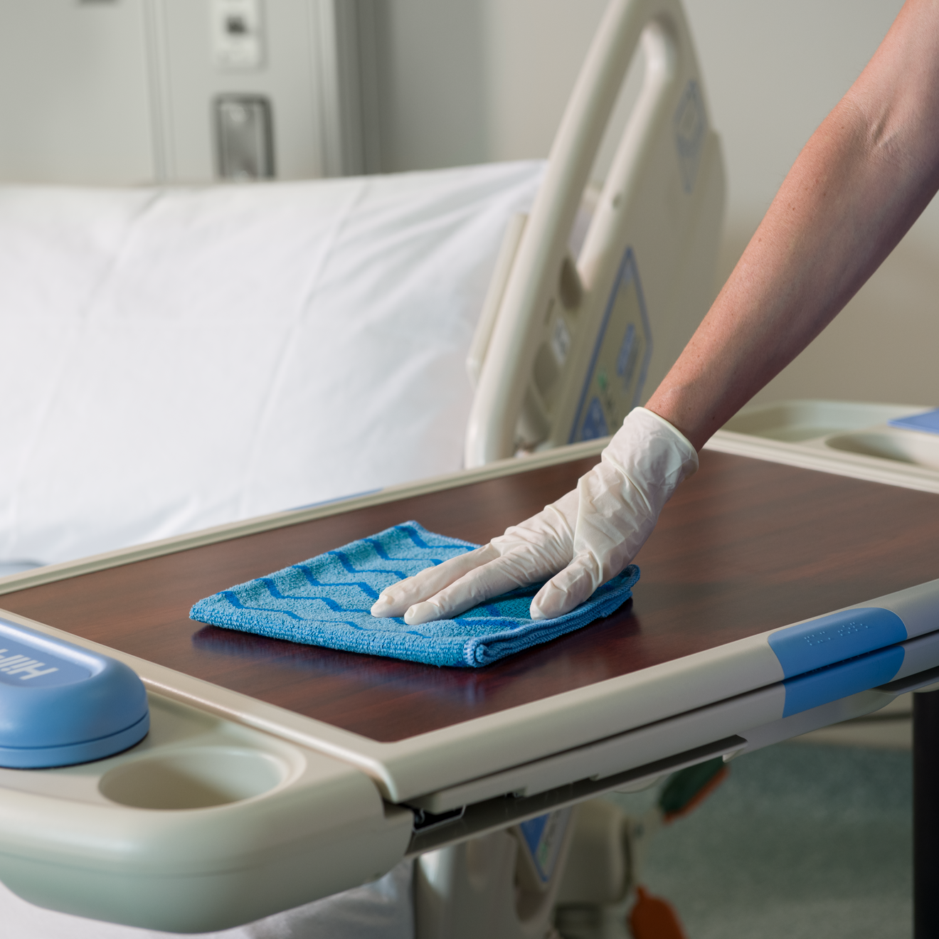 Hand wearing white medical glove cleaning a hospital bedside tray table with a blue Rubbermaid HYGEN™ Microfibre Cloth, with blurred hospital bed in the background.