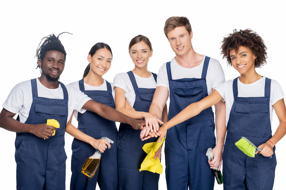 Group of five professional cleaners in uniforms, smiling with hands stacked in teamwork gesture, holding cleaning tools and supplies.